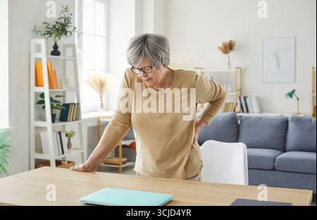Senior woman standing at home touching her back suffering from backache. Stock Photo