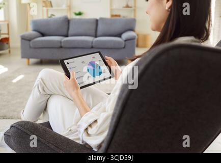 Young woman analyzing financial data at pie chart on tablet screen while working remotely from home. Stock Photo