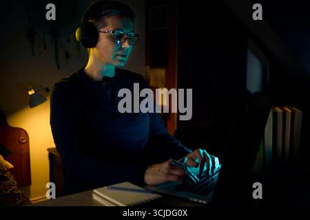 A professional woman with glasses and headphones works late into the evening on her laptop at home. The scene reflects dedication, remote work lifesty Stock Photo