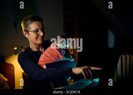 A woman smiles and gestures while working on her laptop with her Canadian Sphynx cat on her lap. The image highlights remote work culture, warmth, and Stock Photo