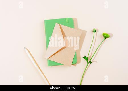 Minimalist flat lay with green notebook, kraft paper envelope, white pencil and green flower on beige background. Top view Stock Photo