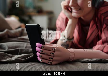 Young adult Caucasian woman with prosthetic hand lying on bed using smartphone, smiling while engaging with device, demonstrating gadget addiction and modern technology interaction Stock Photo