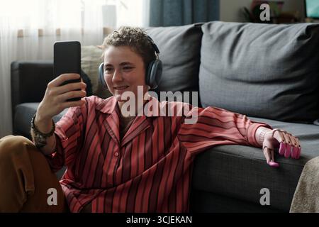 Caucasian teenager sitting on floor using smartphone with headphones, smiling while engaging with device, demonstrating gadget addiction in relaxed home environment Stock Photo