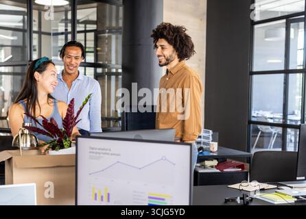 Discussing project with charts, diverse colleagues in modern office using computer Stock Photo