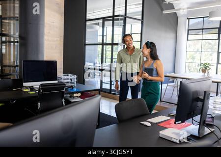 Colleagues in modern office laughing and discussing work near computer desks Stock Photo