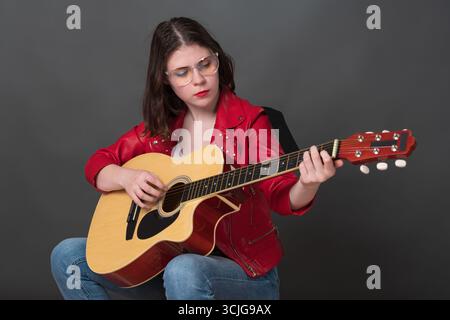 Nice woman is practicing her acoustic guitar and creating warm and lively atmosphere. Young adult female musician, wearing glasses and vibrant red lea Stock Photo