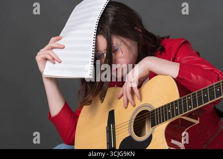 Tired and exhausted female musician with guitar and notes in hand covers face with music sheets due to fatigue from long and tiring rehearsal and lear Stock Photo
