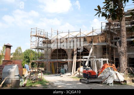 Historic Italian brick building under renovation, surrounded by scaffolding. A mini excavator and construction tools are on-site, with active workers Stock Photo