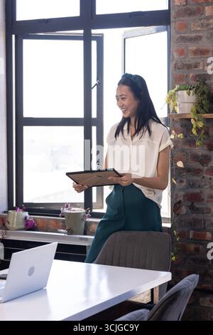 Asian woman holding tablet, smiling in modern office with natural light Stock Photo