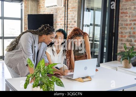 Colleagues collaborating around laptop in modern office, sharing ideas and smiling Stock Photo