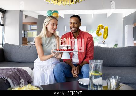 Celebrating birthday, woman presenting cake with candles to smiling man at home Stock Photo