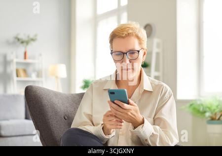 Senior woman in glasses sitting in armchair, using smartphone, enjoying modern technology at home. Stock Photo