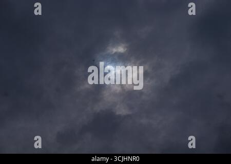 Dramatic hidden sun peeking faintly through thick dark rain clouds in stormy weather Stock Photo