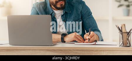 Positive arab male manager working from home during COVID-19 pandemic, using modern laptop and taking notes Stock Photo
