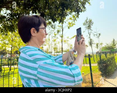 Woman taking a photo outdoors with a smartphone in her hands. Daily life, urban leisure, and personal perspective in an open park environment. Stock Photo