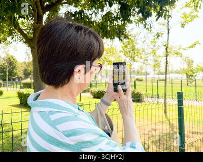 Woman taking a photo outdoors with a smartphone in her hands. Daily life, urban leisure, and personal perspective in an open park environment. Stock Photo