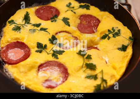 A close-up of a cooked omelette with slices of sausage and fresh cilantro on top. The dish is cooked in a black frying pan Stock Photo