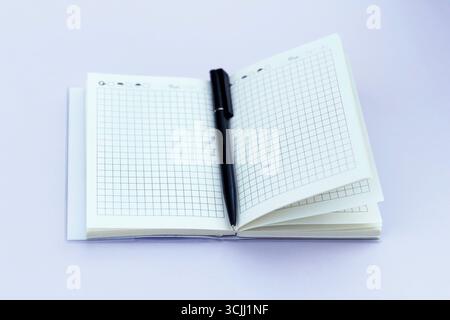 An open notebook with a black pen resting on a blank page. Notebook features grid paper and is placed on a light purple background Stock Photo