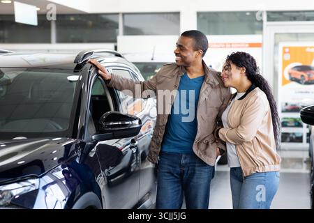 Young African American couple smiling while discussing new SUV inside dealership showroom Stock Photo
