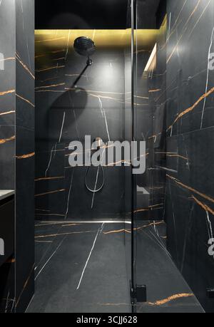 Empty shower room with dark grey stone tiles with orange veins. Opened glass door in stylish shower interior design in dark tones, warm illumination. Stock Photo