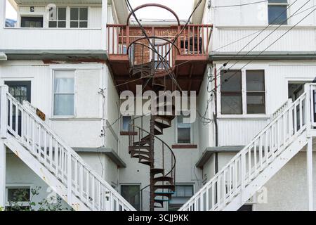 Spiral staircase is seen in front of a building with a white exterior. The staircase is old and rusted, and it is located on the side of the building. Stock Photo