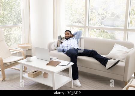 Relaxed smiling young man recline on comfy sofa using phone Stock Photo