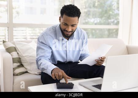 Busy smiling young man using calculator to do budgeting work Stock Photo