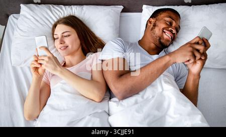 Gadget addiction and relationship problems. Millennial interracial couple with smartphones lying in bed, top view Stock Photo