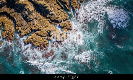 Ocean waves hitting rugged rocky shore seen from above with foamy white water and clear blue sea powerful coastal landscape and nature scenery Stock Photo