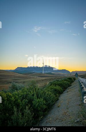 Empty asphalt road leading through Spanish countryside with wind turbines and mountains at sunset in Navarra region Stock Photo