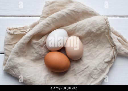 Top down view of three different chicken eggs — white, brown, and beige — resting on a linen napkin placed on a white wooden surface. Rustic farm fres Stock Photo
