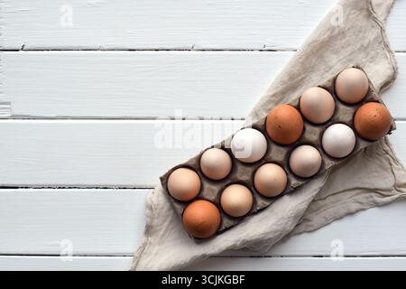 Top down view of a carton with a dozen assorted chicken eggs in various sizes and colors placed on a white wooden surface with a linen napkin and copy Stock Photo