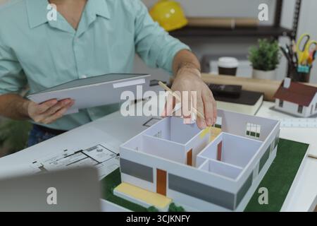Asian male architect is analyzing a modern house model while holding the roof structure and pointing at layout with a pencil. Concept of architectural Stock Photo