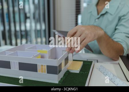 Asian male architect is analyzing a modern house model while holding the roof structure and pointing at layout with a pencil. Concept of architectural Stock Photo