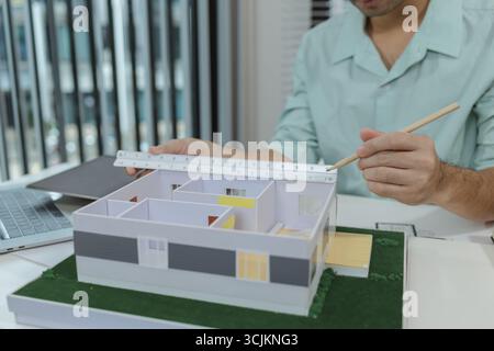 Asian male architect sitting at a desk, measuring a modern house model with triangular ruler and pencil. Office setup includes design tools, blueprint Stock Photo