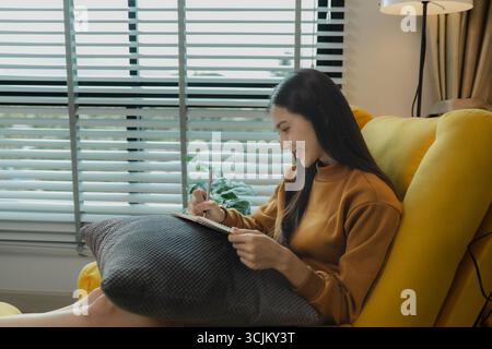An Asian woman is sitting on the sofa in the living room. She is keeping a diary to record past memories and experiences. Keeping a diary is a leisure Stock Photo