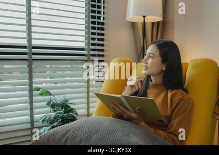 An Asian woman is sitting on the sofa in the living room. She is keeping a diary to record past memories and experiences. Keeping a diary is a leisure Stock Photo