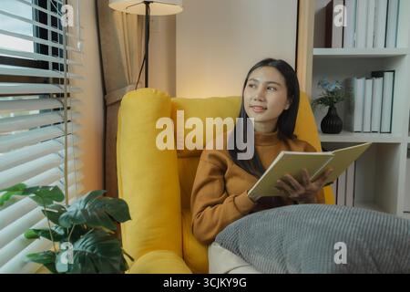 An Asian woman is sitting on the sofa in the living room. She is keeping a diary to record past memories and experiences. Keeping a diary is a leisure Stock Photo