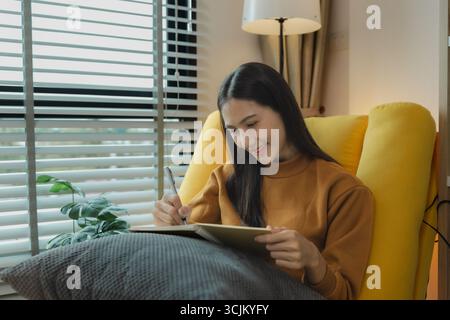 An Asian woman is sitting on the sofa in the living room. She is keeping a diary to record past memories and experiences. Keeping a diary is a leisure Stock Photo