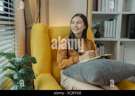 An Asian woman is sitting on the sofa in the living room. She is keeping a diary to record past memories and experiences. Keeping a diary is a leisure Stock Photo