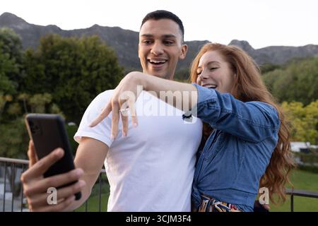 Young couple taking selfie outdoors, showing engagement ring with joyful expressions Stock Photo