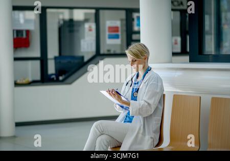 Young female doctor with short hair in sitting on bench in hospital corridor, reviewing documentation. Stock Photo