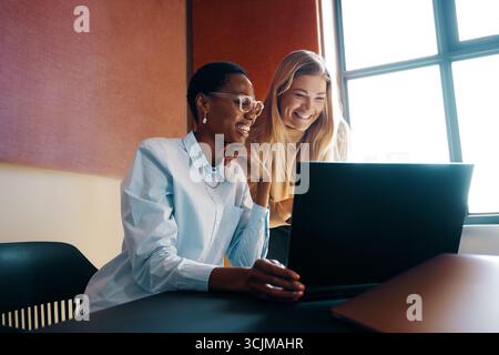 Two young women, one African and one caucasian, collaborating and smiling while sharing ideas in a modern office environment. Stock Photo