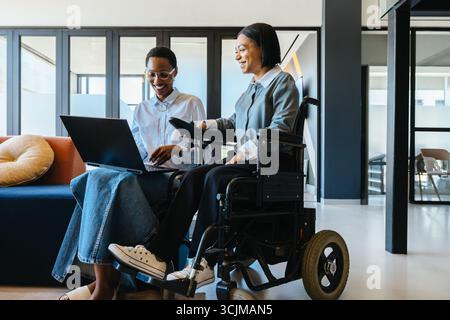 Two women, one a wheelchair user, are collaborating in a modern office setting, using a laptop. Stock Photo