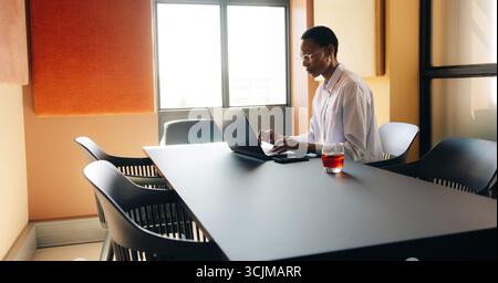 A young African woman using a laptop in a modern office setting, natural lighting highlights the scene. The environment conveys focus and productivity Stock Photo