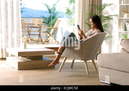 Happy woman using mobile phone sitting on armchair at home Stock Photo