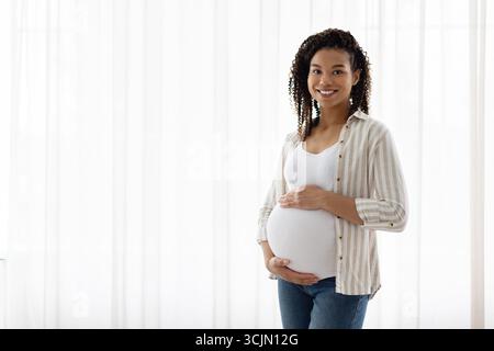 Smiling pregnant woman stands by white curtains with a hand on belly Stock Photo