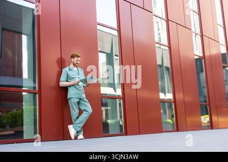 Doctor in scrubs standing by wall reading from open medical folder Stock Photo