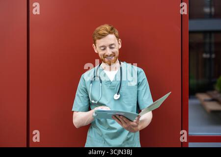 Man doctor holding open paper folder while leaning on wall Stock Photo