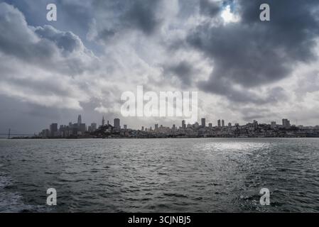 View of the distant cityscape under a dramatic sky with the sun reflecting on the water in a moody atmosphere, San Francisco, California, United States. Stock Photo